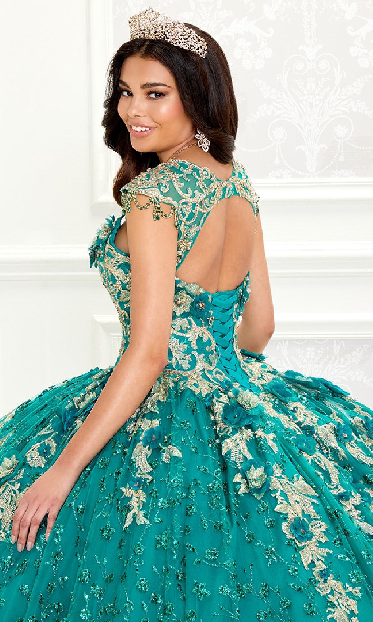A woman wearing the Quinceanera Dress PR30088 by Princesa in ornate teal and gold with lace embroidery and a tiara stands in a decorated room, looking over her shoulder.